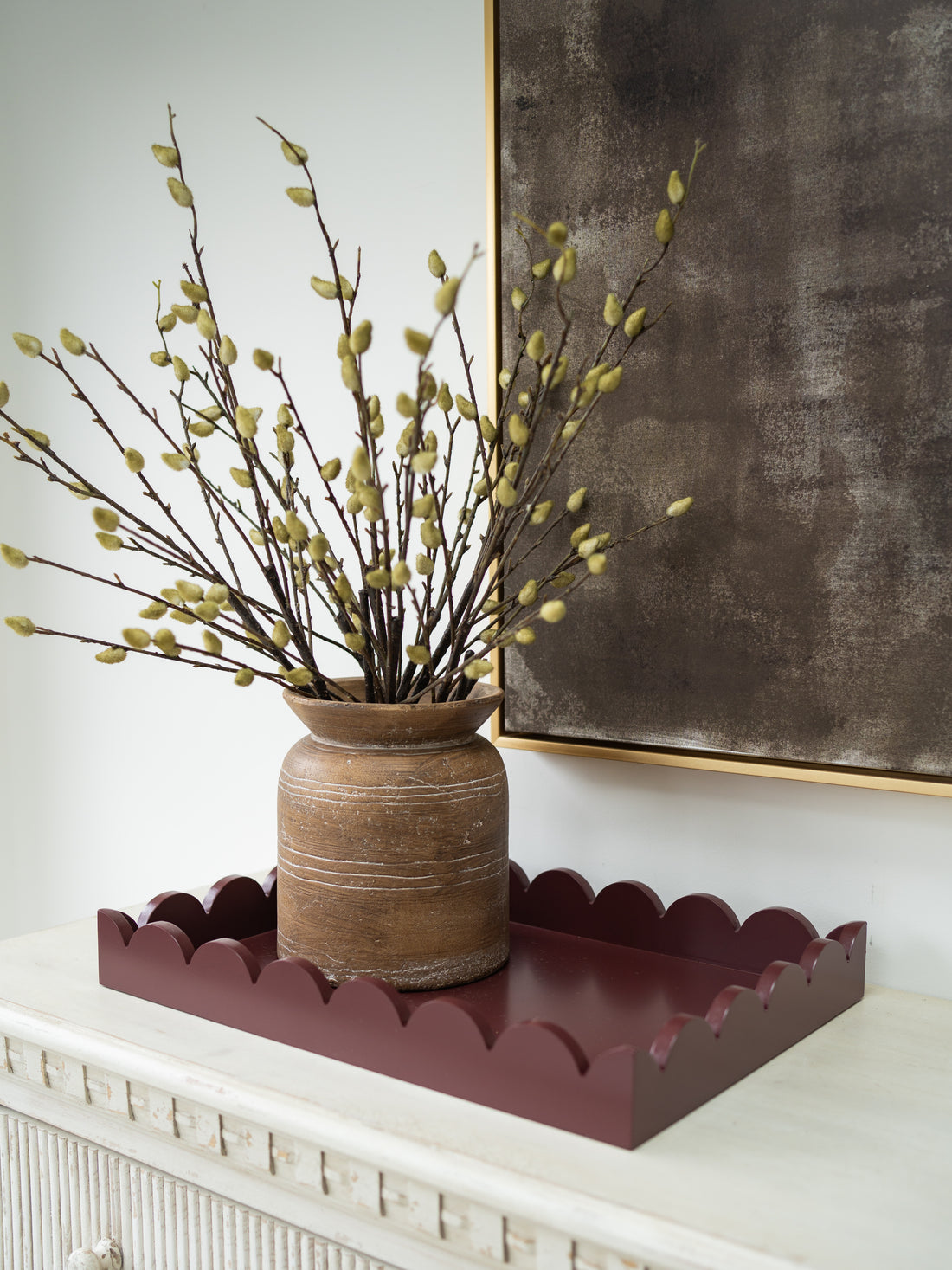 A rustic brown ceramic vase filled with willow branches with light green buds, displayed on a scalloped dark red tray atop a white console table, with a dark textured wall and gold-framed art in the background.