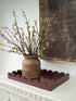 A rustic brown ceramic vase filled with willow branches with light green buds, displayed on a scalloped dark red tray atop a white console table, with a dark textured wall and gold-framed art in the background.