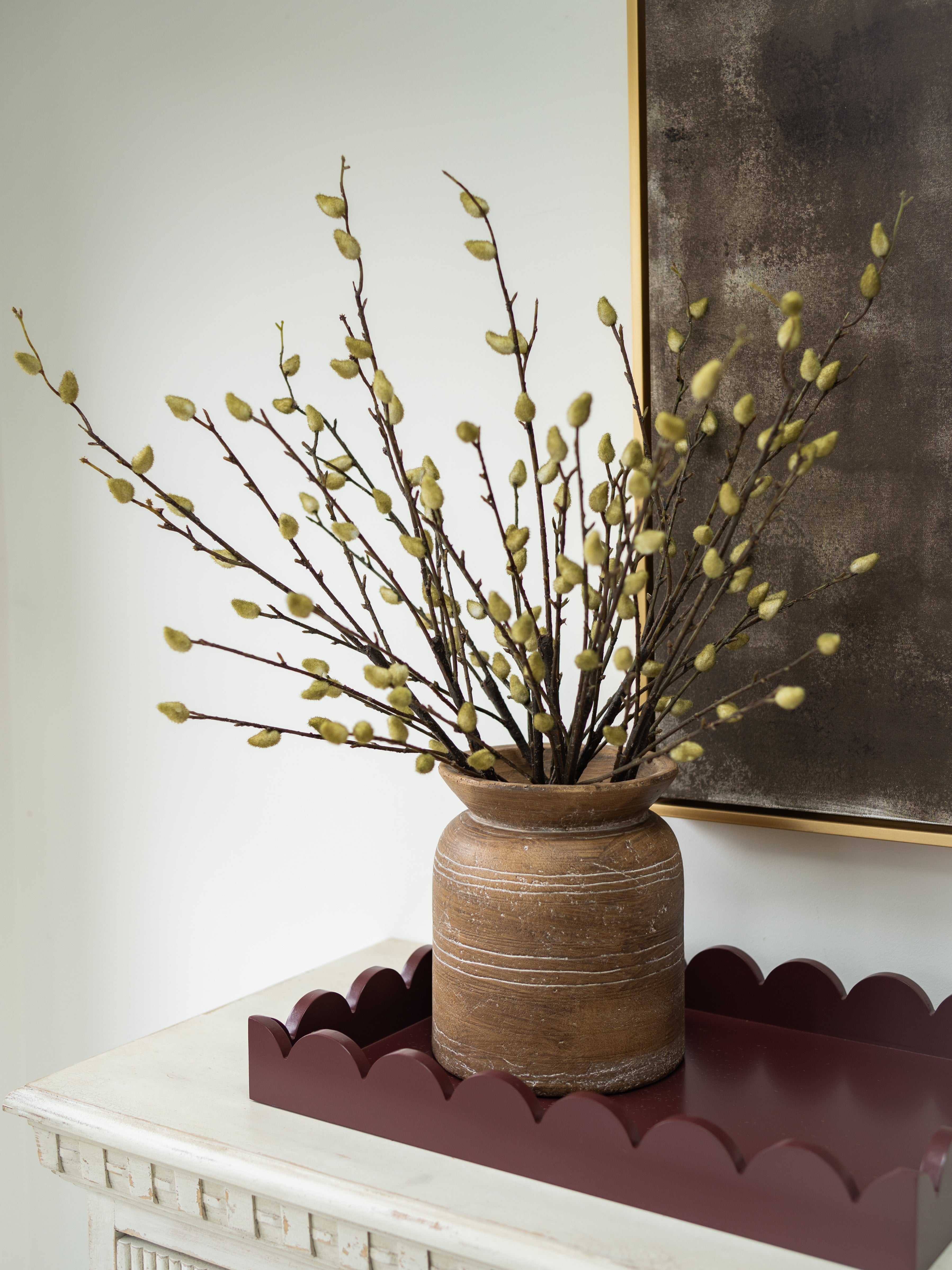 A rustic brown ceramic vase filled with faux pussy willow branches featuring light green buds, displayed on a scalloped dark red tray atop a white console table against a white wall and a dark framed artwork.