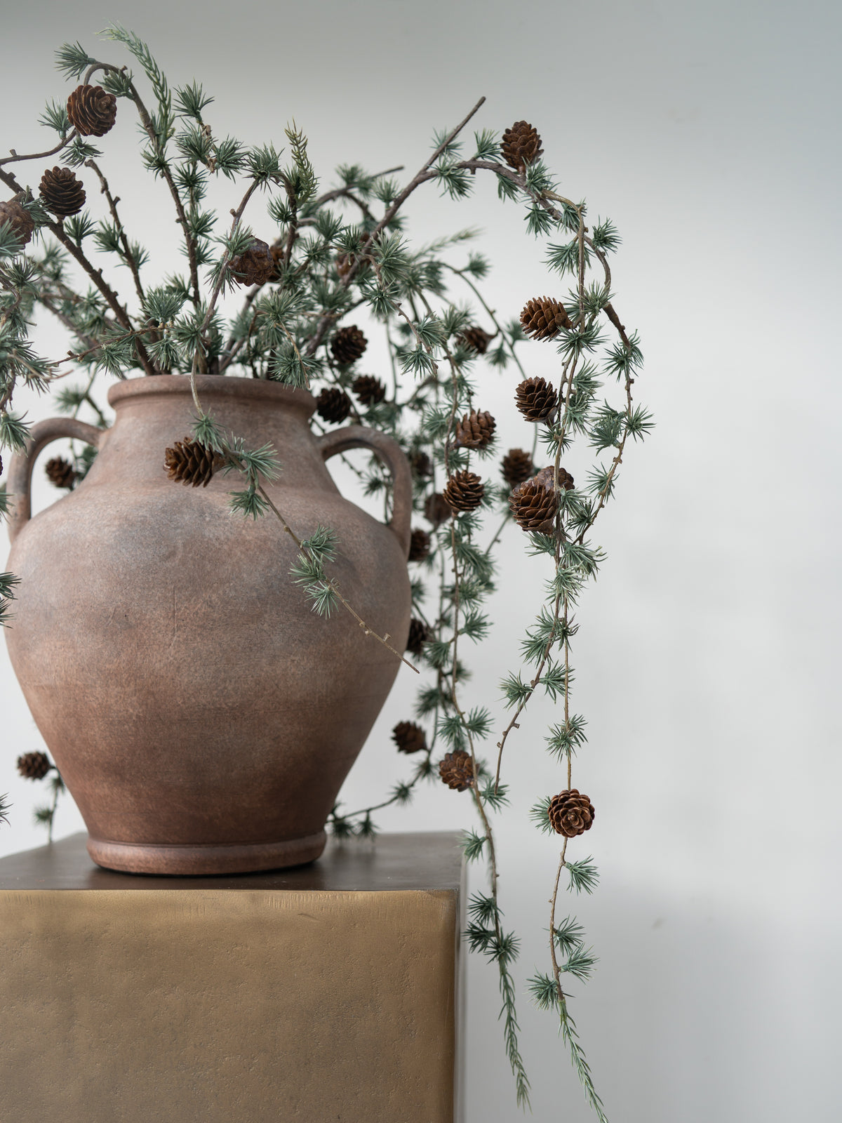 A rustic brown ceramic vase with two handles, filled with pine branches and pinecones, displayed on a gold-toned pedestal against a light gray background.