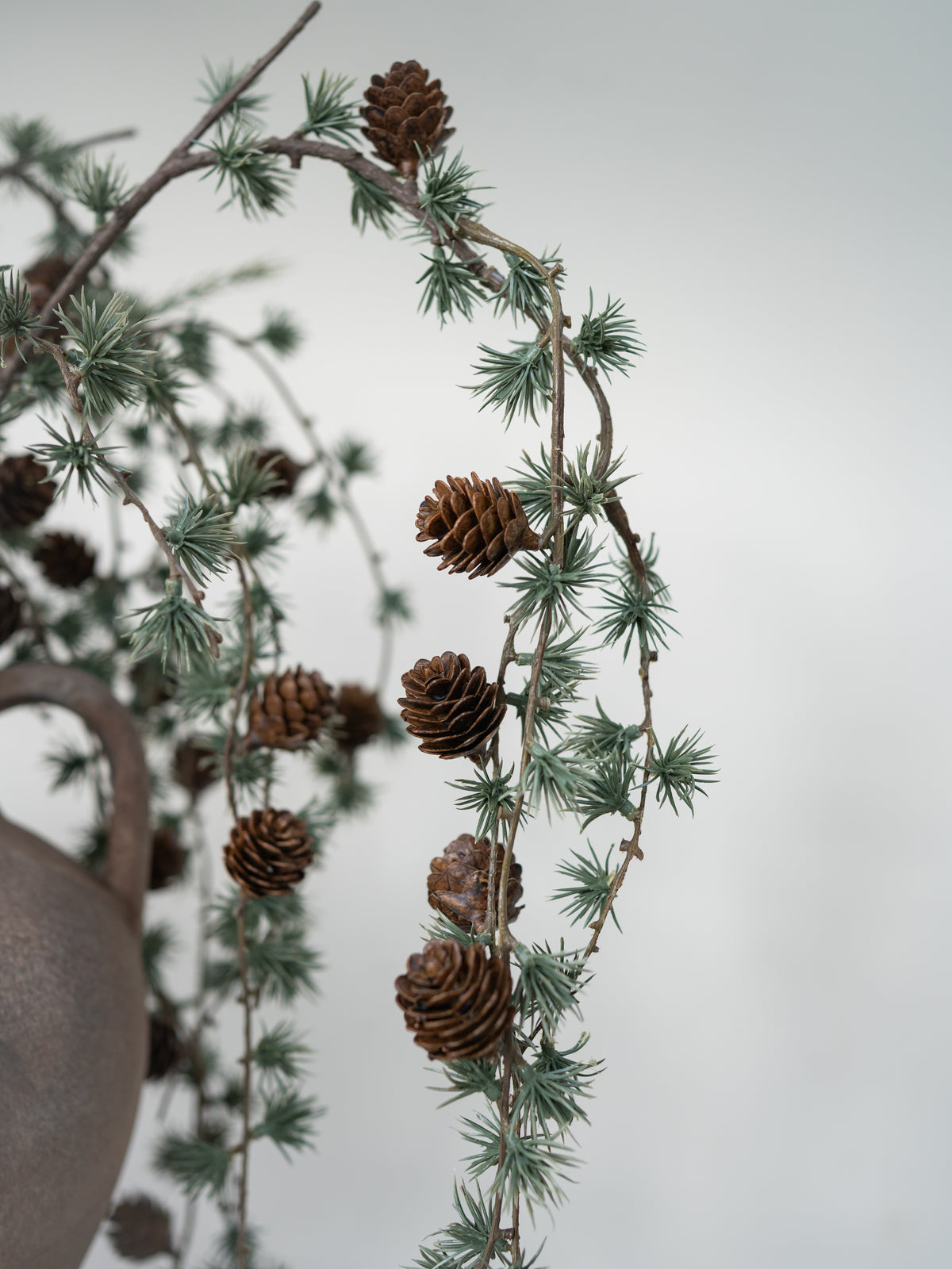 Pinecones on a pine branch with green needles, arranged decoratively with a rustic ceramic pot partially visible on the left against a soft light gray background.
