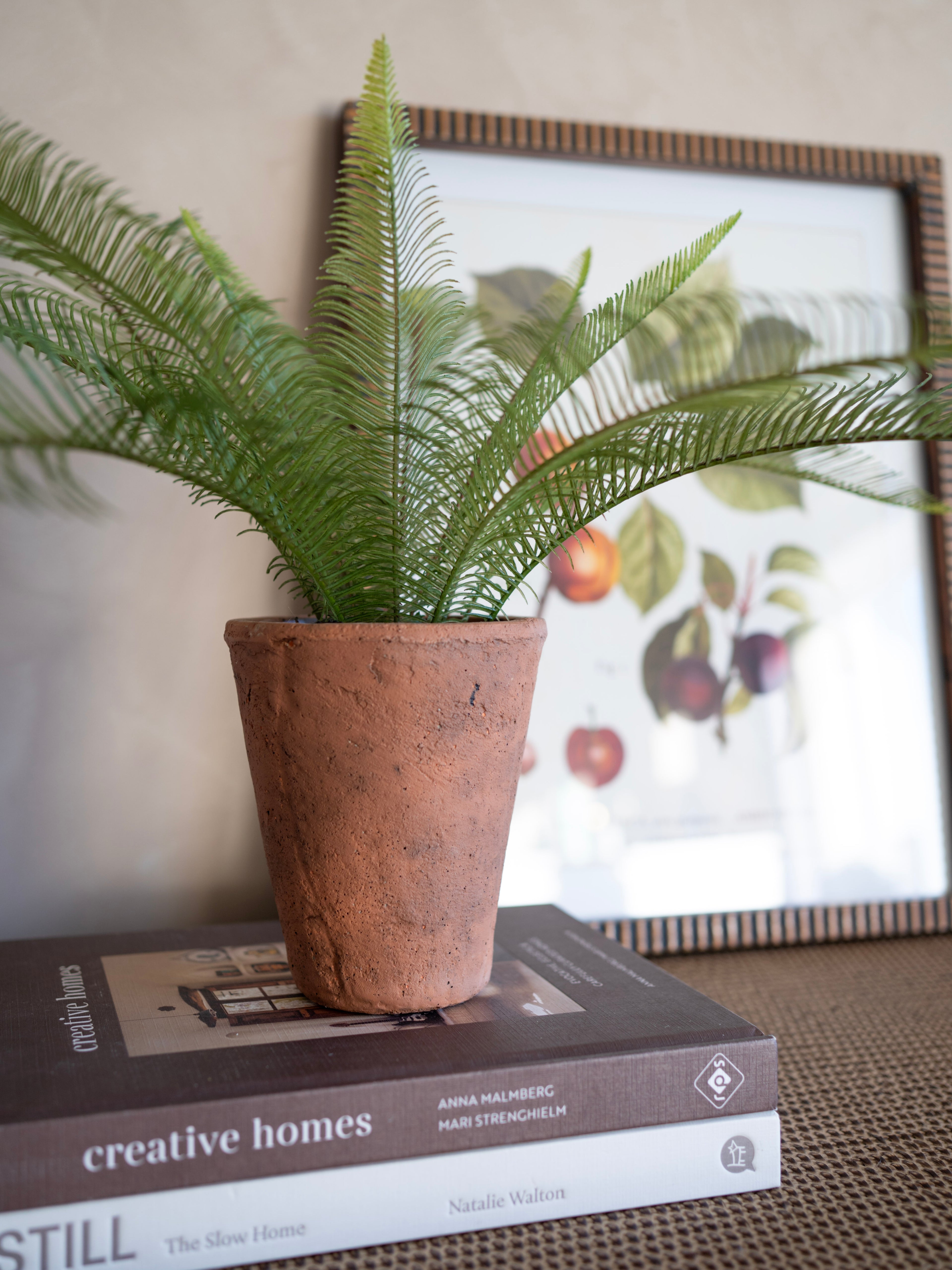 Potted faux java fern plant on top of books with a blurred artwork in the background