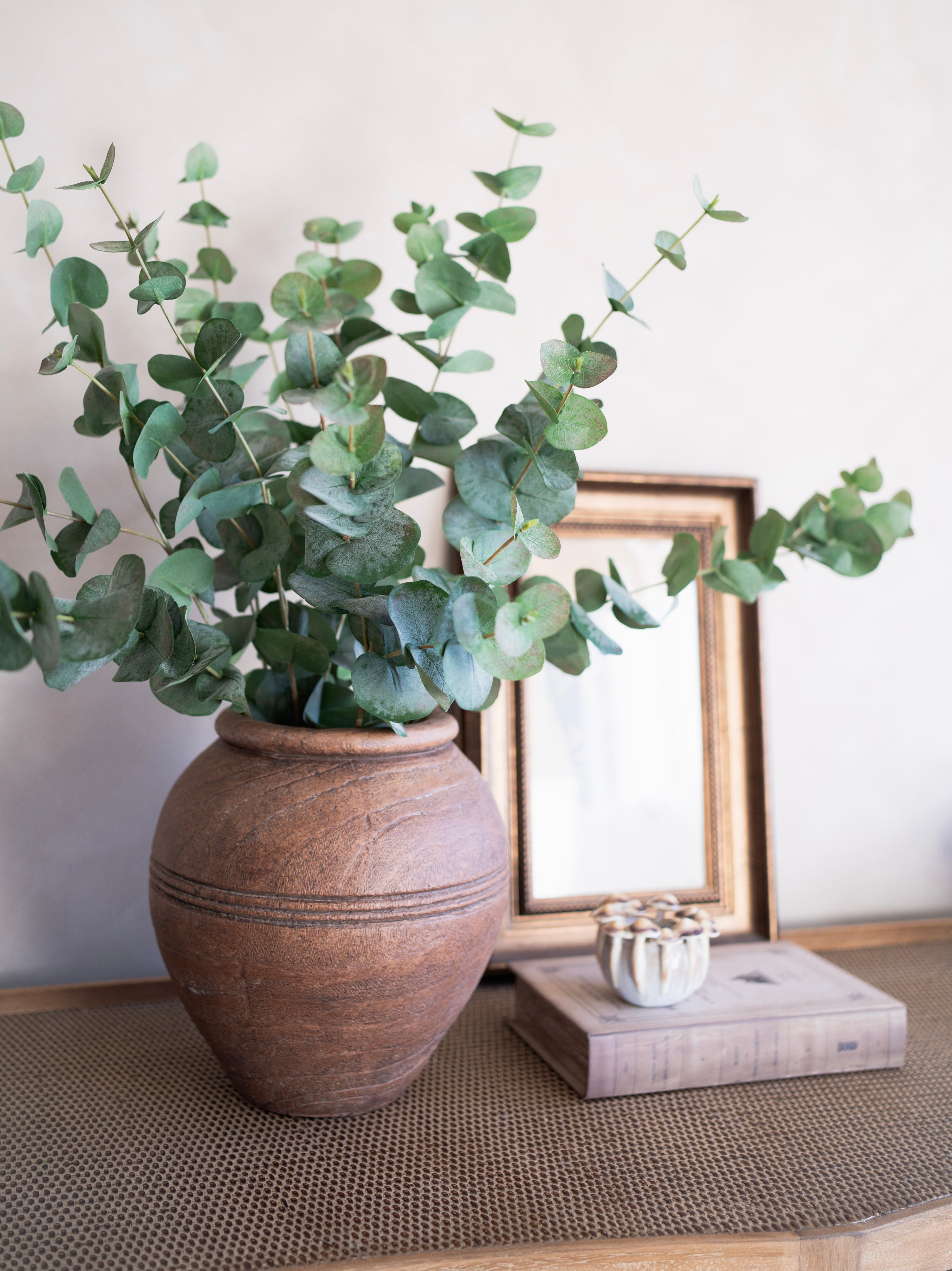 faux green eucalyptus in a terracotta pot on a wooden surface with a framed picture in the background.
