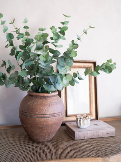faux green eucalyptus in a terracotta pot on a wooden surface with a framed picture in the background.