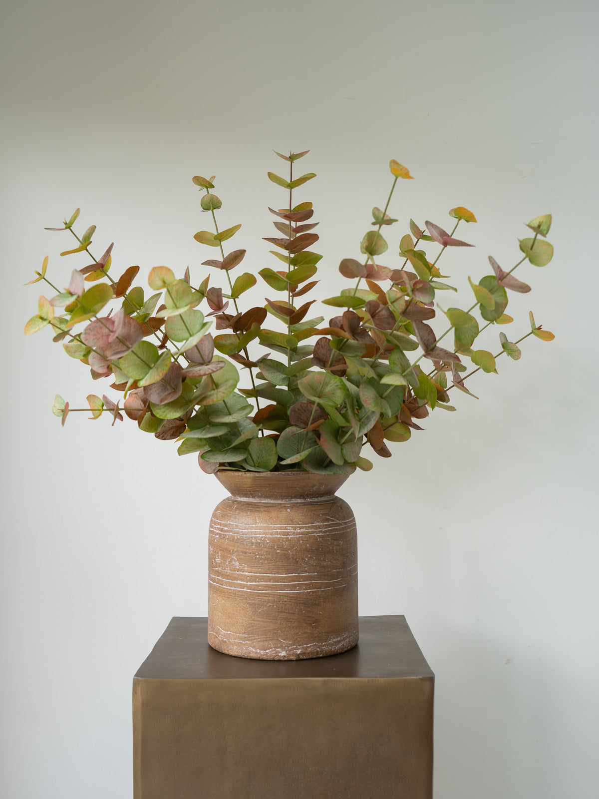 A faux eucalyptus floral arrangement with green and brown leaves in a rustic terracotta pot, displayed on a dark bronze pedestal against a light gray wall.