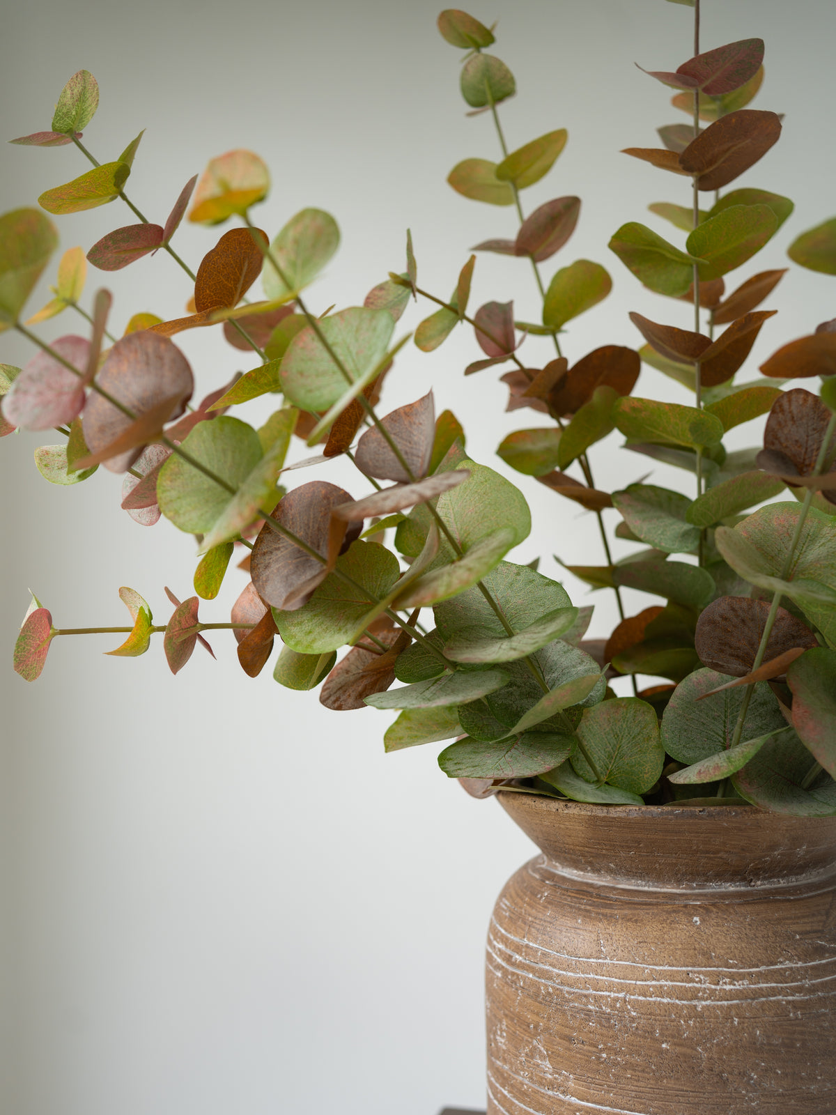 Artificial eucalyptus branch with green and brownish leaves arranged in a rustic brown ceramic vase against a plain light gray background.