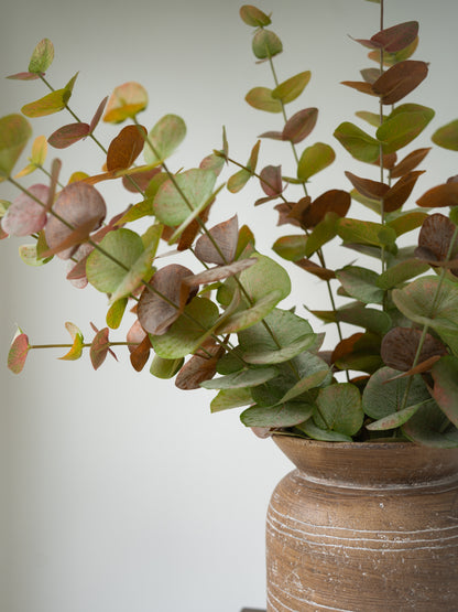 Artificial eucalyptus branch with green and brownish leaves arranged in a rustic brown ceramic vase against a plain light gray background.