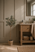 Faux Natural Olive Tree in Aged Pot in bathroom setting with wooden sink unit with brass taps, baskets with hand towels and bathroom mat in the foreground