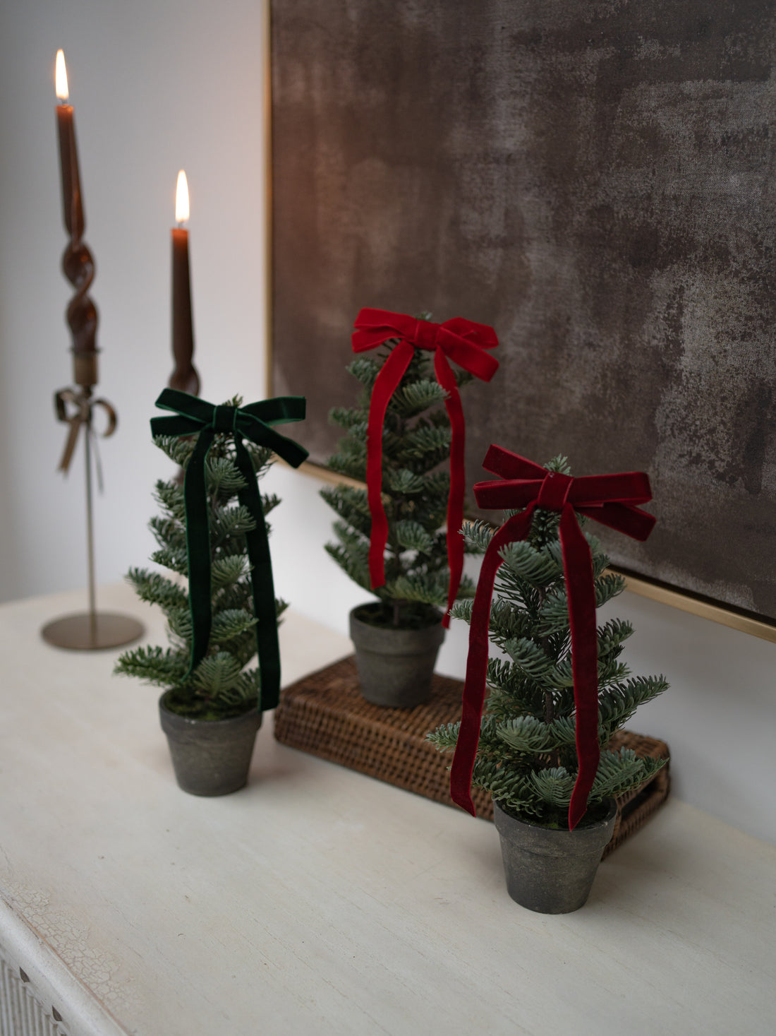 Three small artificial Christmas tree toppers in gray pots, decorated with dark green and red velvet ribbon bows, arranged on a white mantel with lit candles and a textured wall in the background.