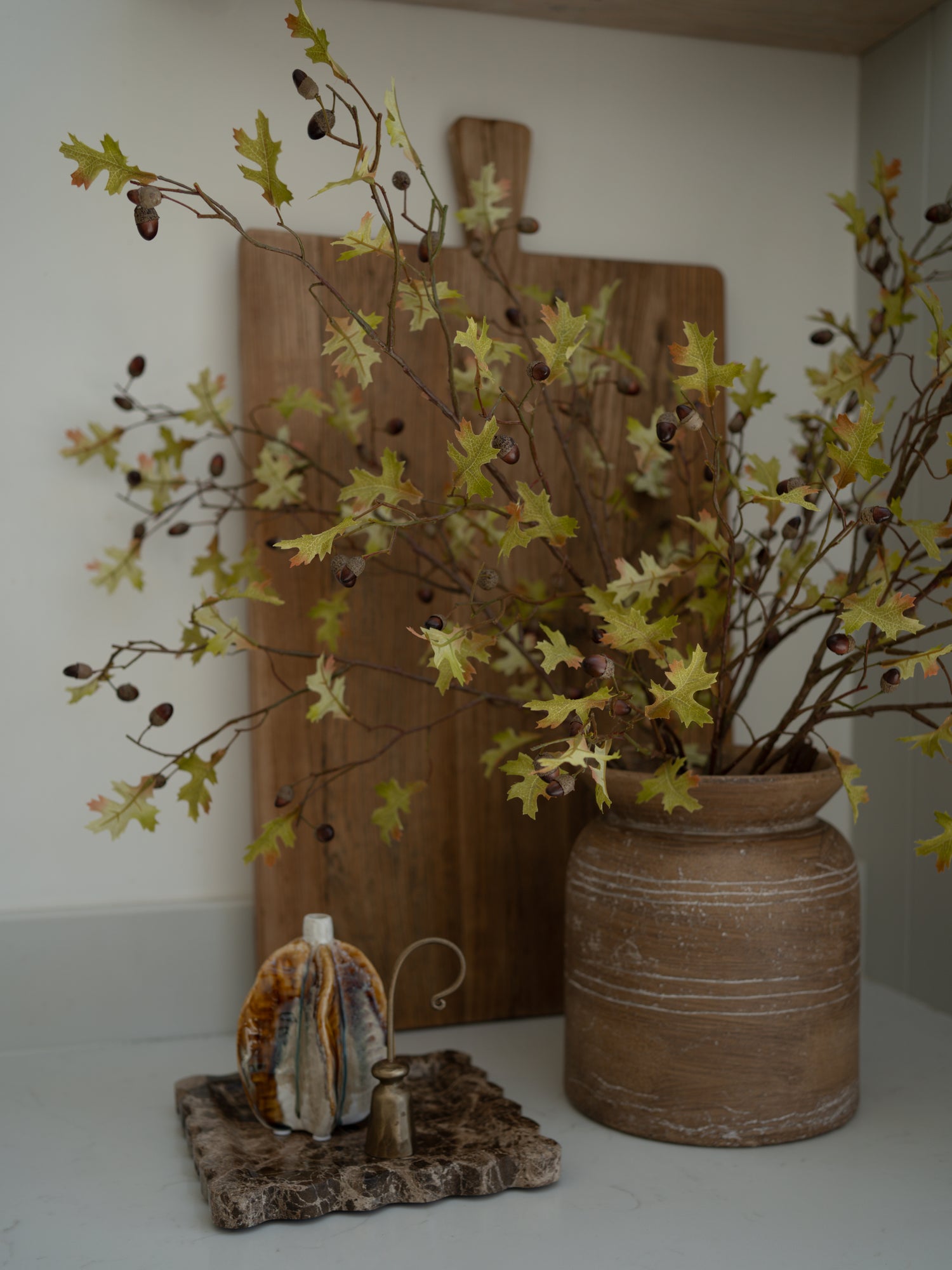Decorative setup with a vase of faux oak and acorn branches, wooden cutting board, and small decorative item on a white surface.