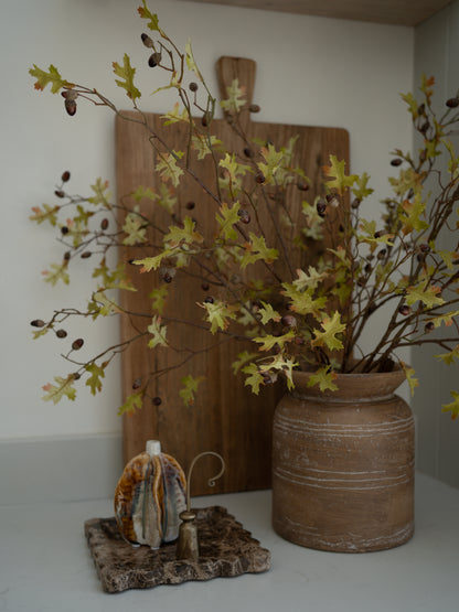Decorative setup with a vase of faux oak and acorn branches, wooden cutting board, and small decorative item on a white surface.
