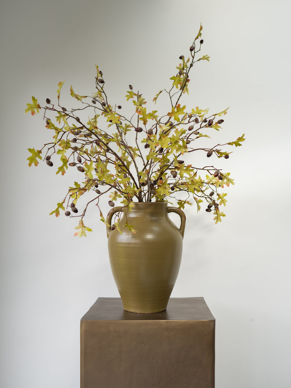 An artificial autumn leaf and acorn branch arrangement in a brown ceramic vase with two side handles, displayed on a square pedestal against a plain light gray background.