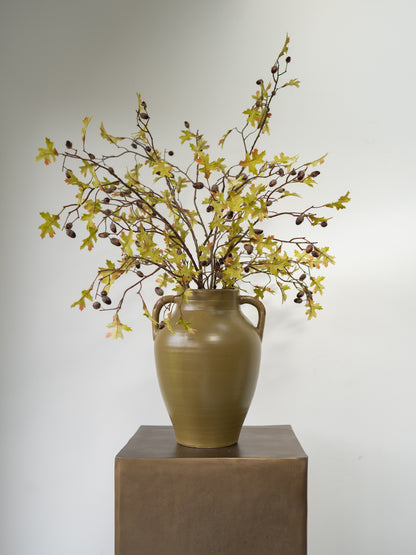 An artificial autumn leaf and acorn branch arrangement in a brown ceramic vase with two side handles, displayed on a square pedestal against a plain light gray background.