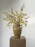 An artificial autumn leaf and acorn branch arrangement in a brown ceramic vase with two side handles, displayed on a square pedestal against a plain light gray background.