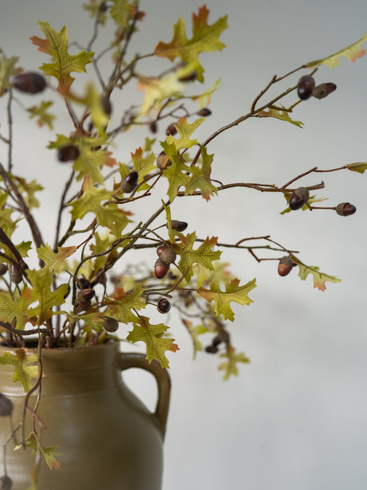Autumn oak branch arrangement with green and yellow leaves, brown acorns, and brown twigs, displayed in a matte brown ceramic vase against a soft light gray background.