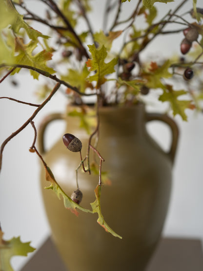 Autumn oak leaf branch arrangement with acorns in a matte olive-green ceramic vase, photographed against a soft light gray background.