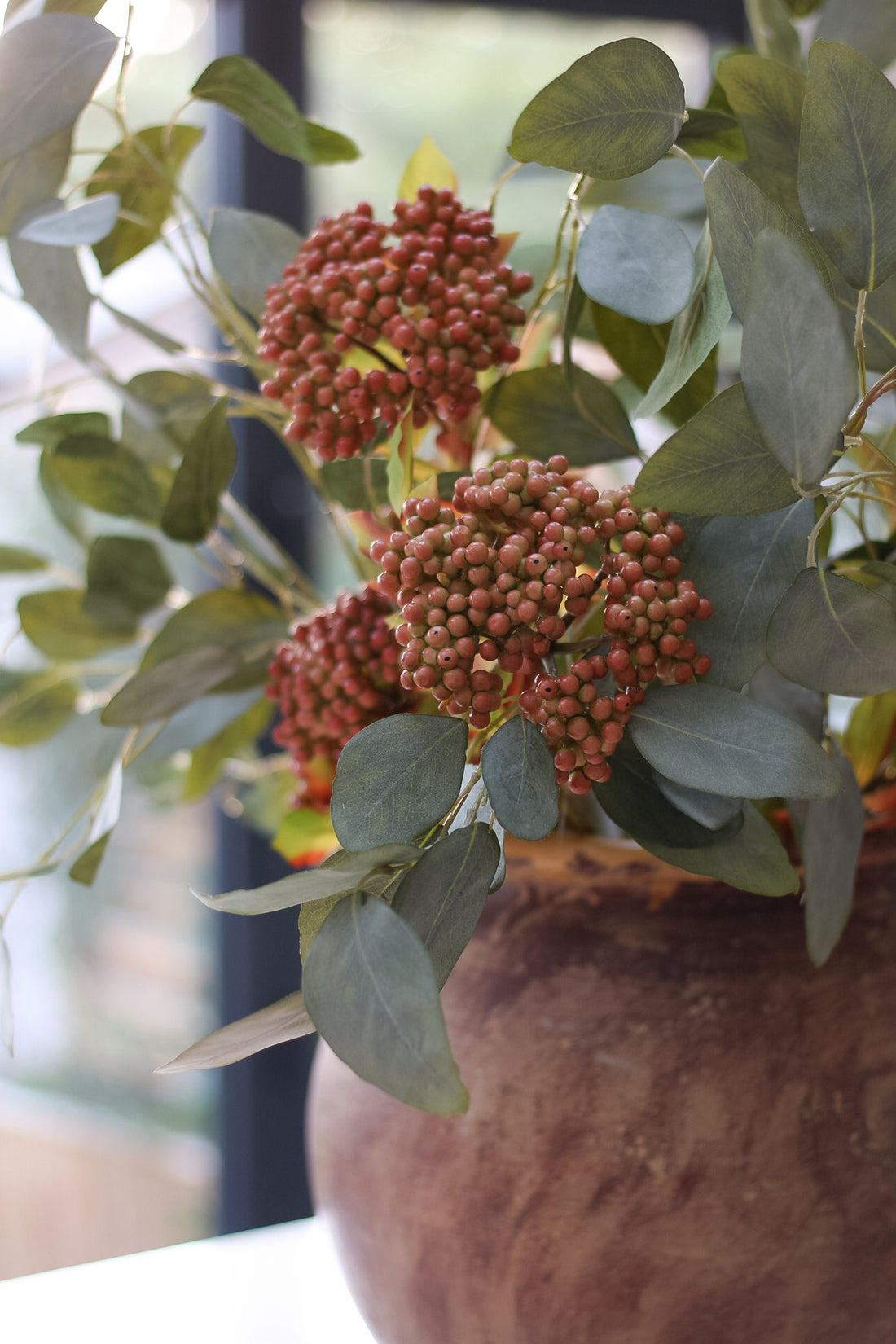 Decorative arrangement with faux orange berries and green leaves in a brown pot.