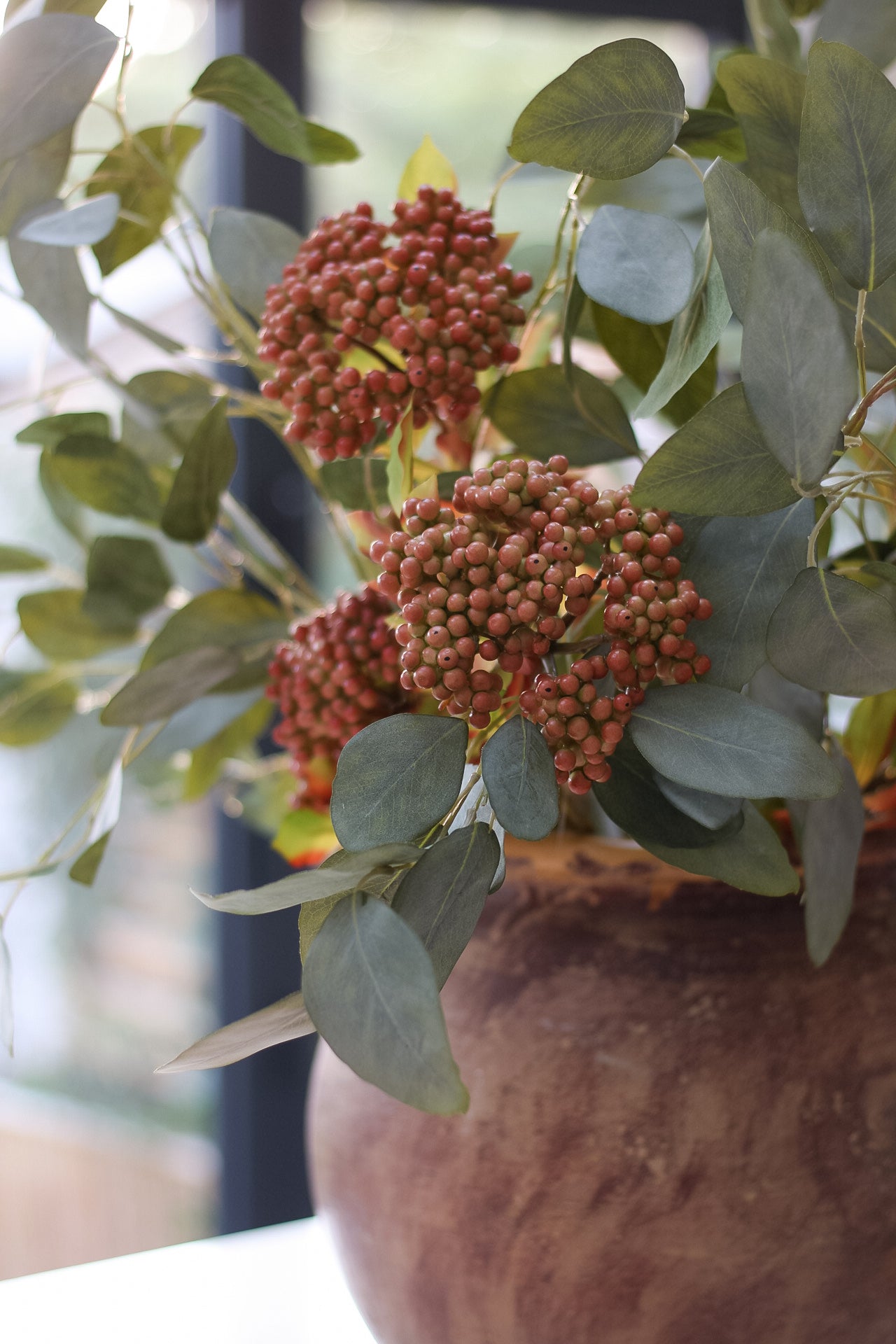 Decorative arrangement with faux orange berries and green leaves in a brown pot.