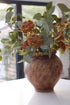 Decorative vase with faux eucalyptus green leaves and orange berries on a white surface