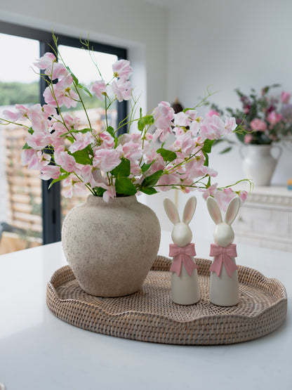 Decorative setting with a vase of pink flowers, two white rabbit figurines with pink bows, and a woven tray on a table.