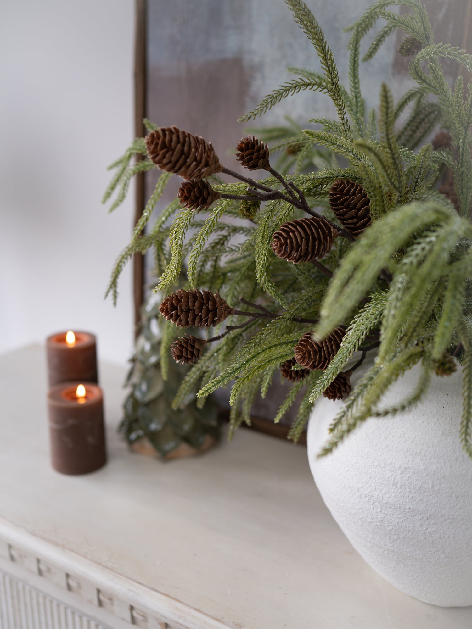 Decorative setup with a vase of greenery, candles, and pinecones on a white surface.