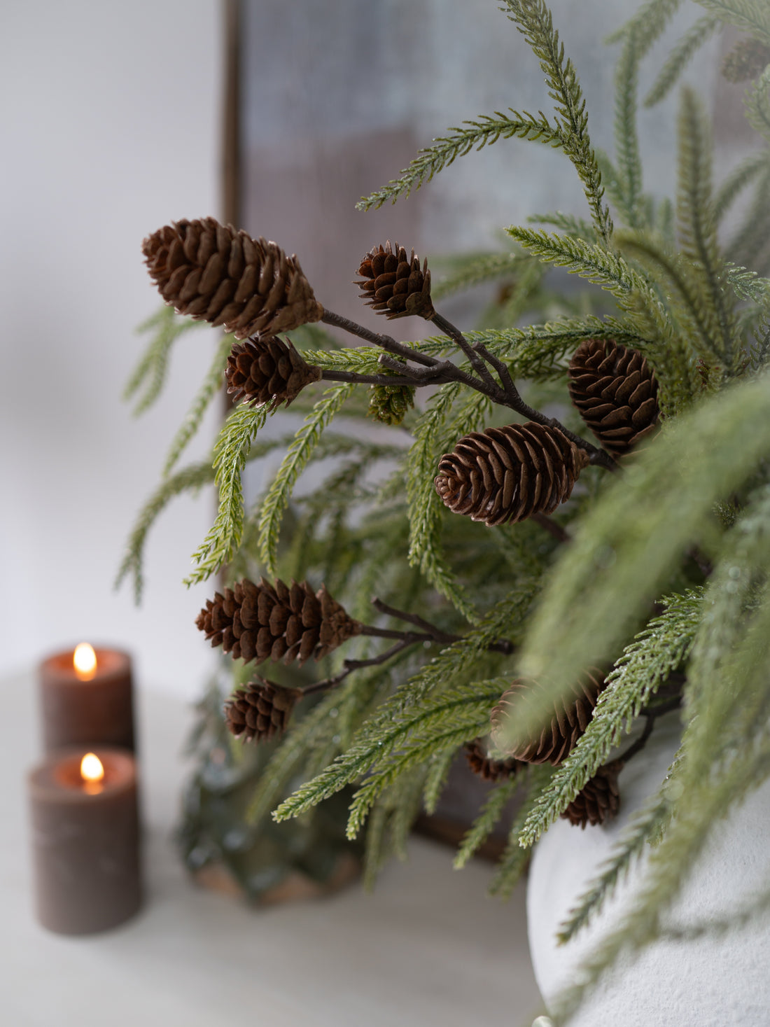 Decorative arrangement with greenery and pine cones on a white surface with candles.