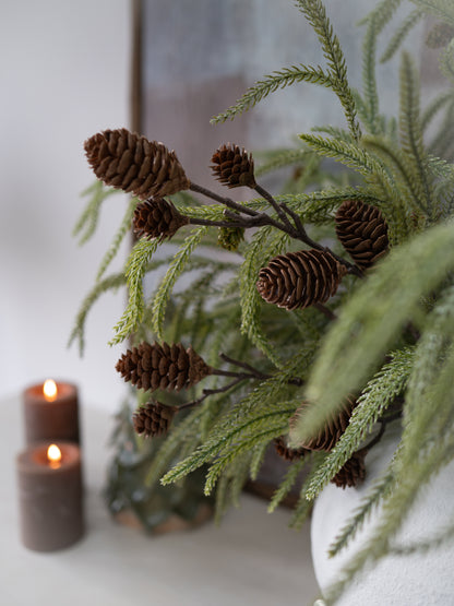 Decorative arrangement with greenery and pine cones on a white surface with candles.