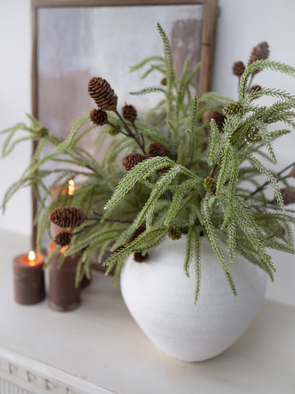 White vase with green foliage and pinecones on a white surface with candles and a framed picture in the background.