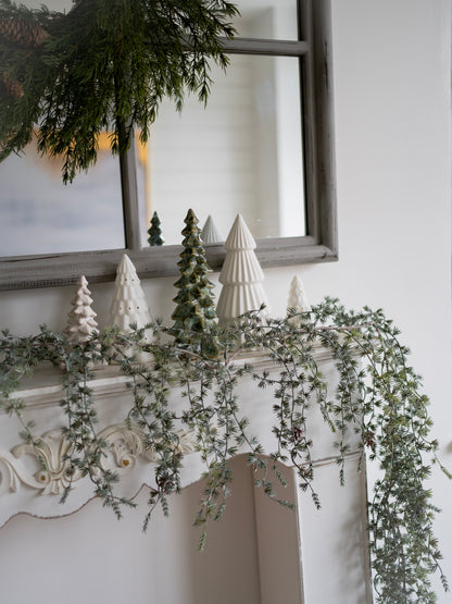 Decorative mantel with greenery, white ceramic trees, and a mirror in the background.