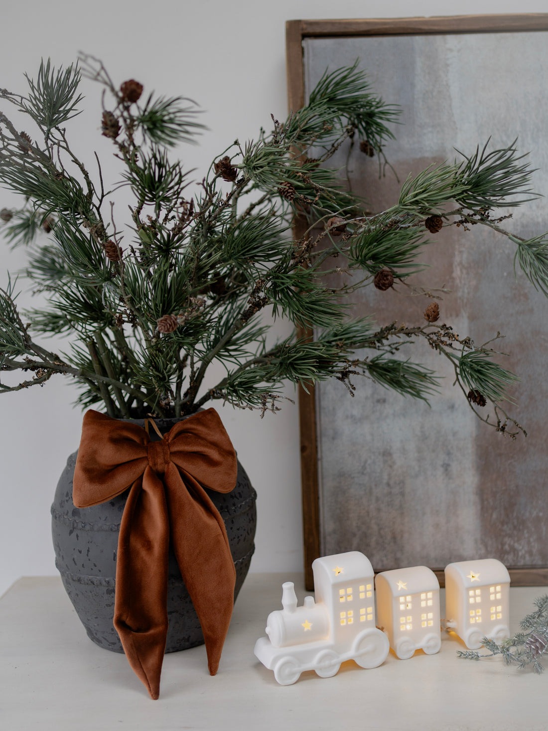 Decorative arrangement with a vase of greenery, brown bow, and small illuminated houses on a white surface.