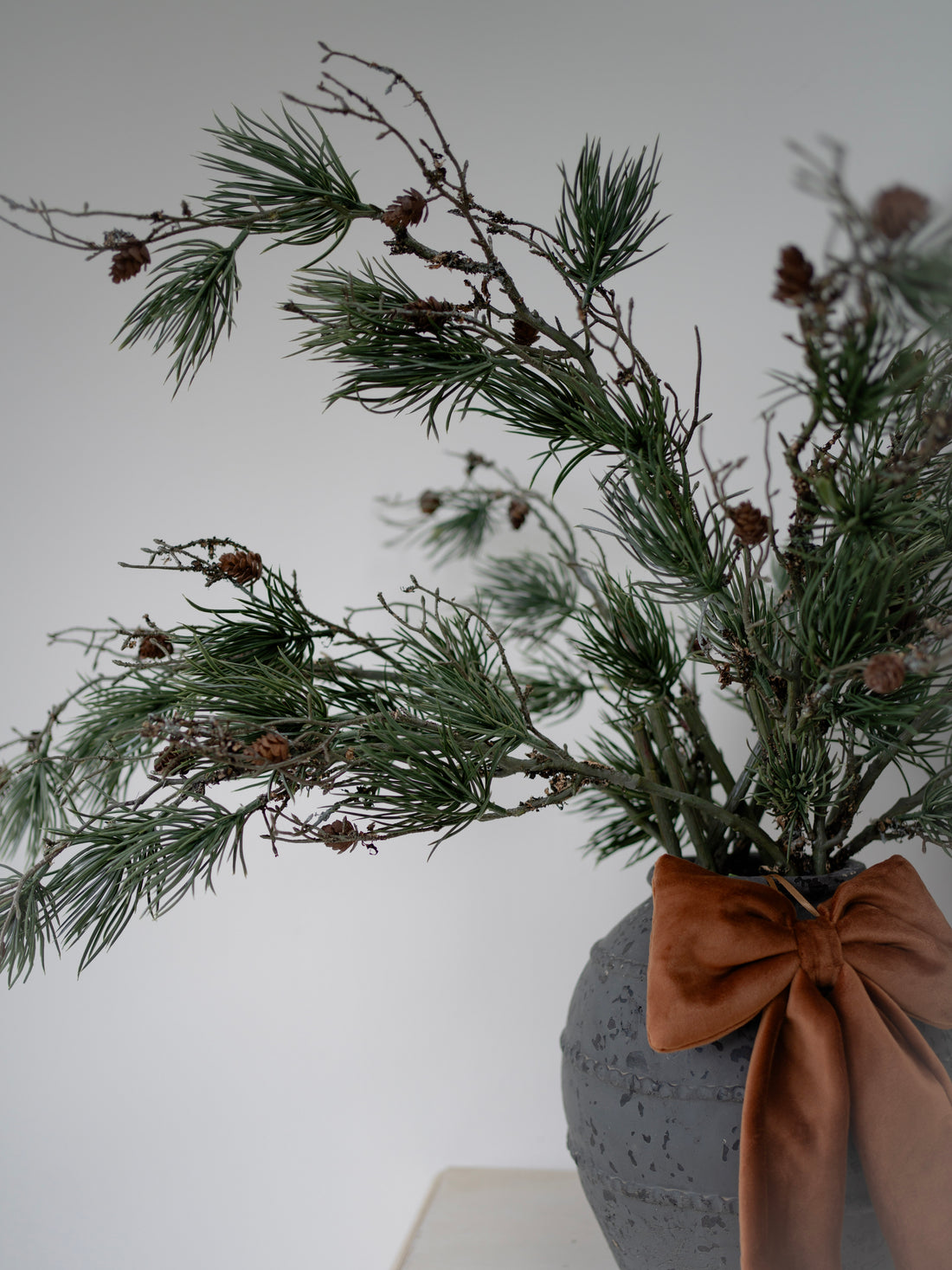 Decorative arrangement of greenery with pinecones in a textured gray vase on a white surface.