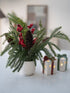 Decorative arrangement with greenery, red berries, and pinecones in a pot on a table with candles and gift boxes.