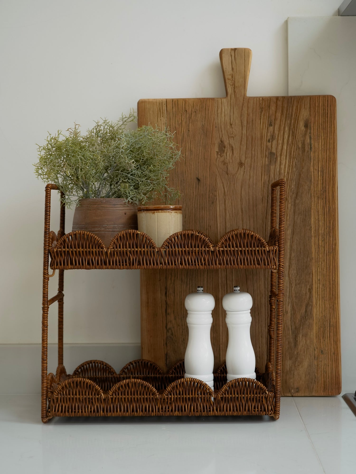 Wicker shelf with decorative items including a wooden cutting board, plants, and salt and pepper shakers on a light background.