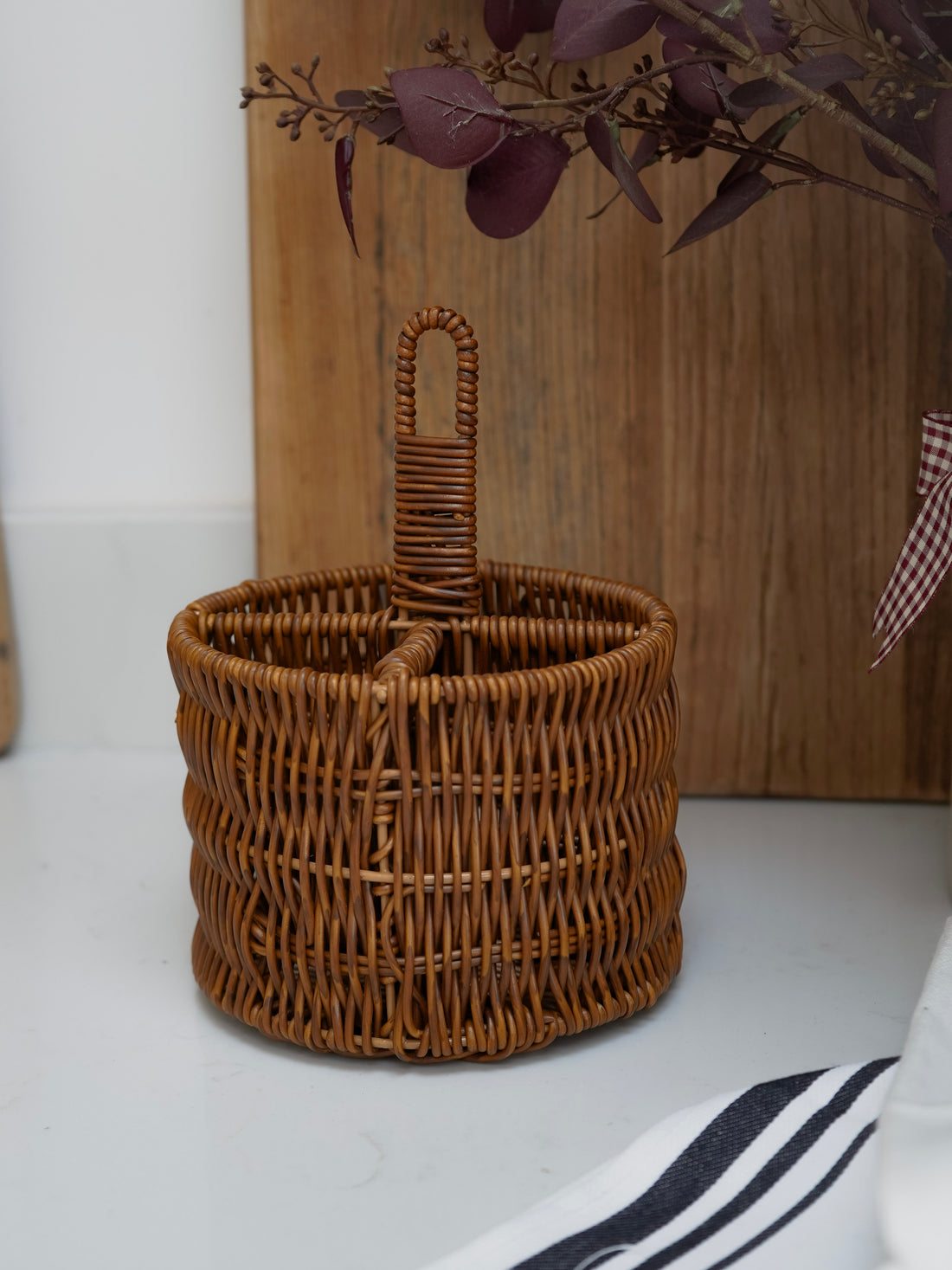Wicker utensil basket with handle on a white surface with a wooden board and plant in the background