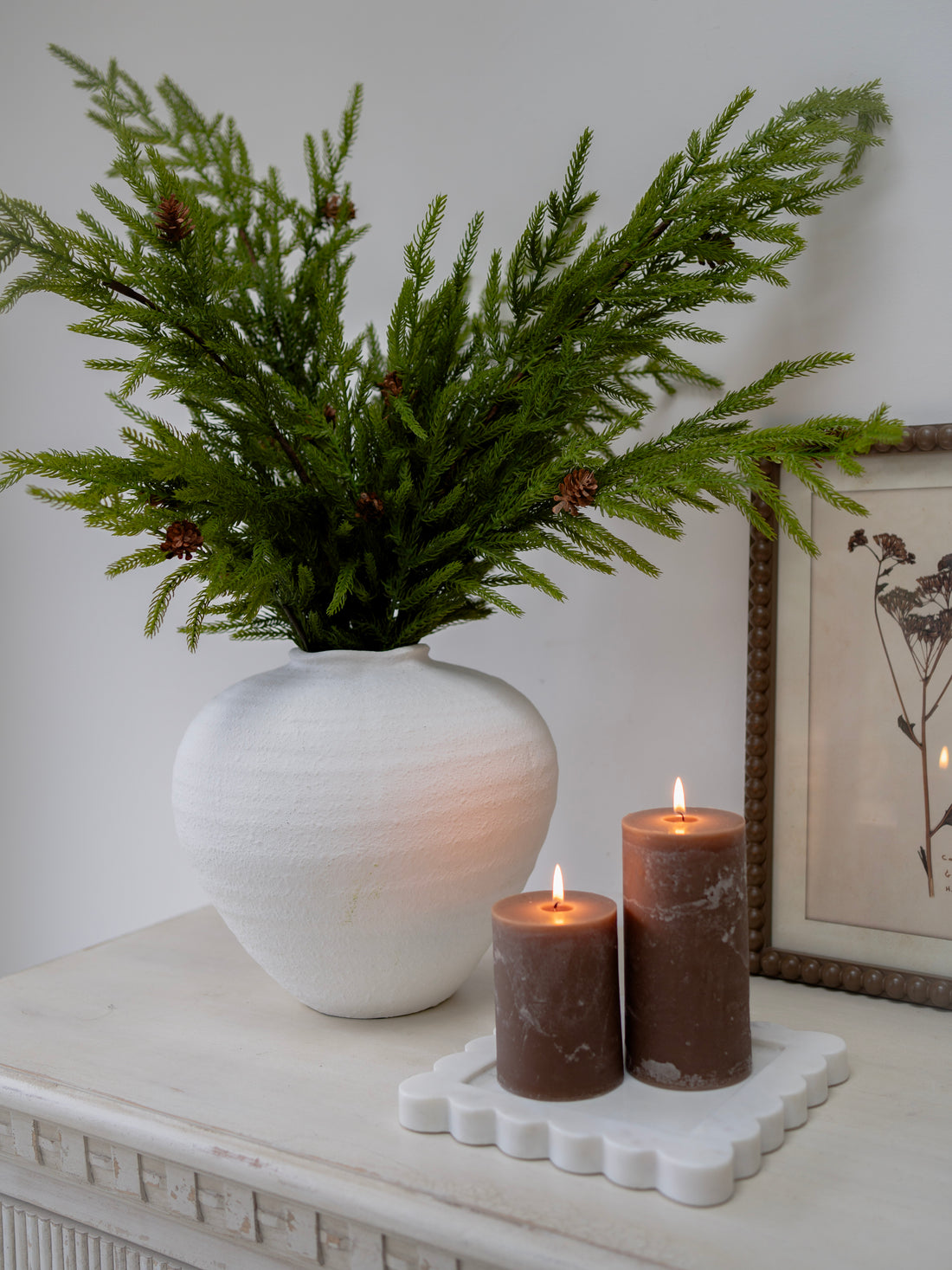 White vase with faux norfolk pine greenery, two brown candles on a white surface with a framed picture in the background.