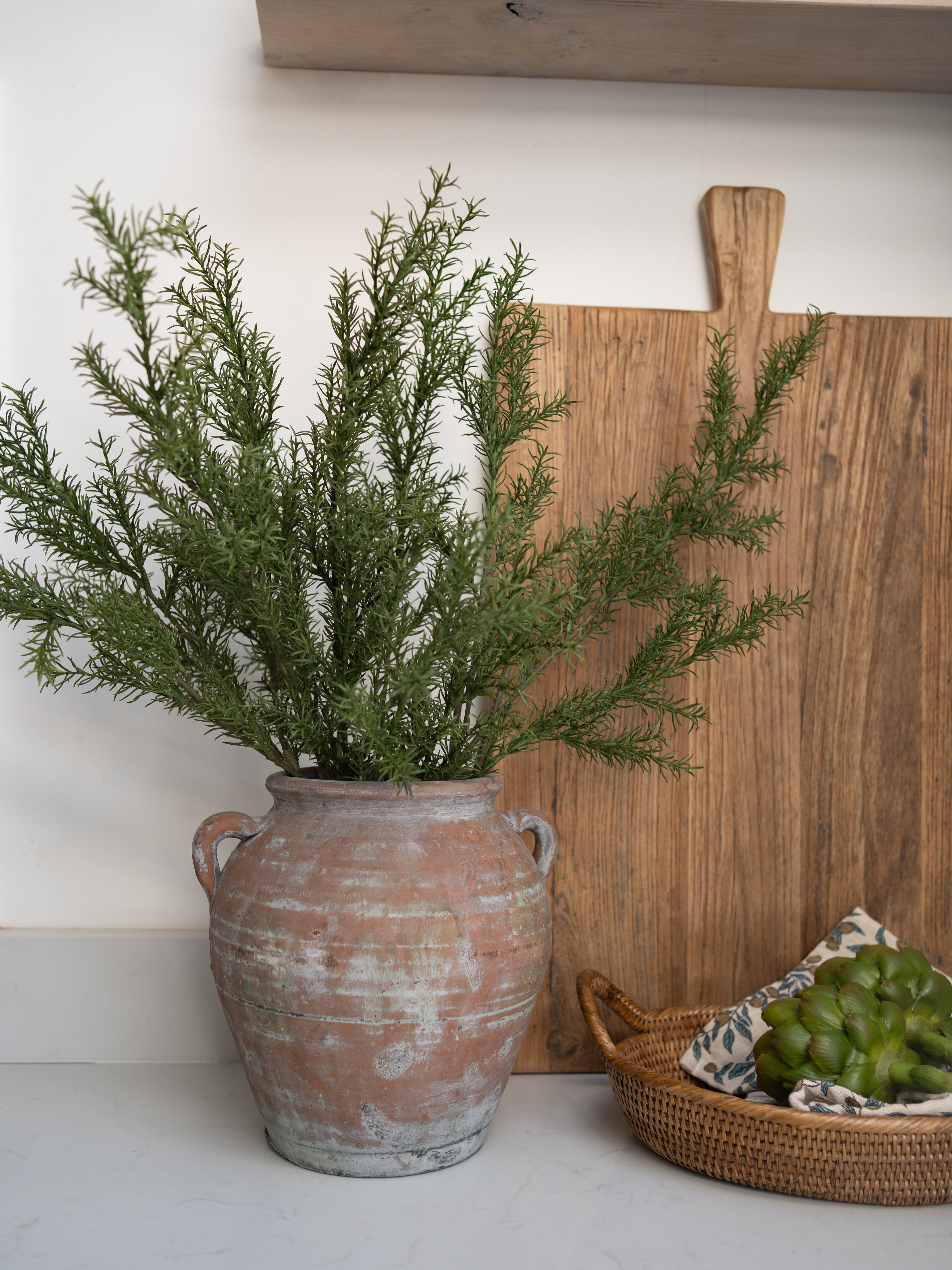 Faux rosemary stems in a rustic pot on a white surface with a wooden cutting board and basket in the background.