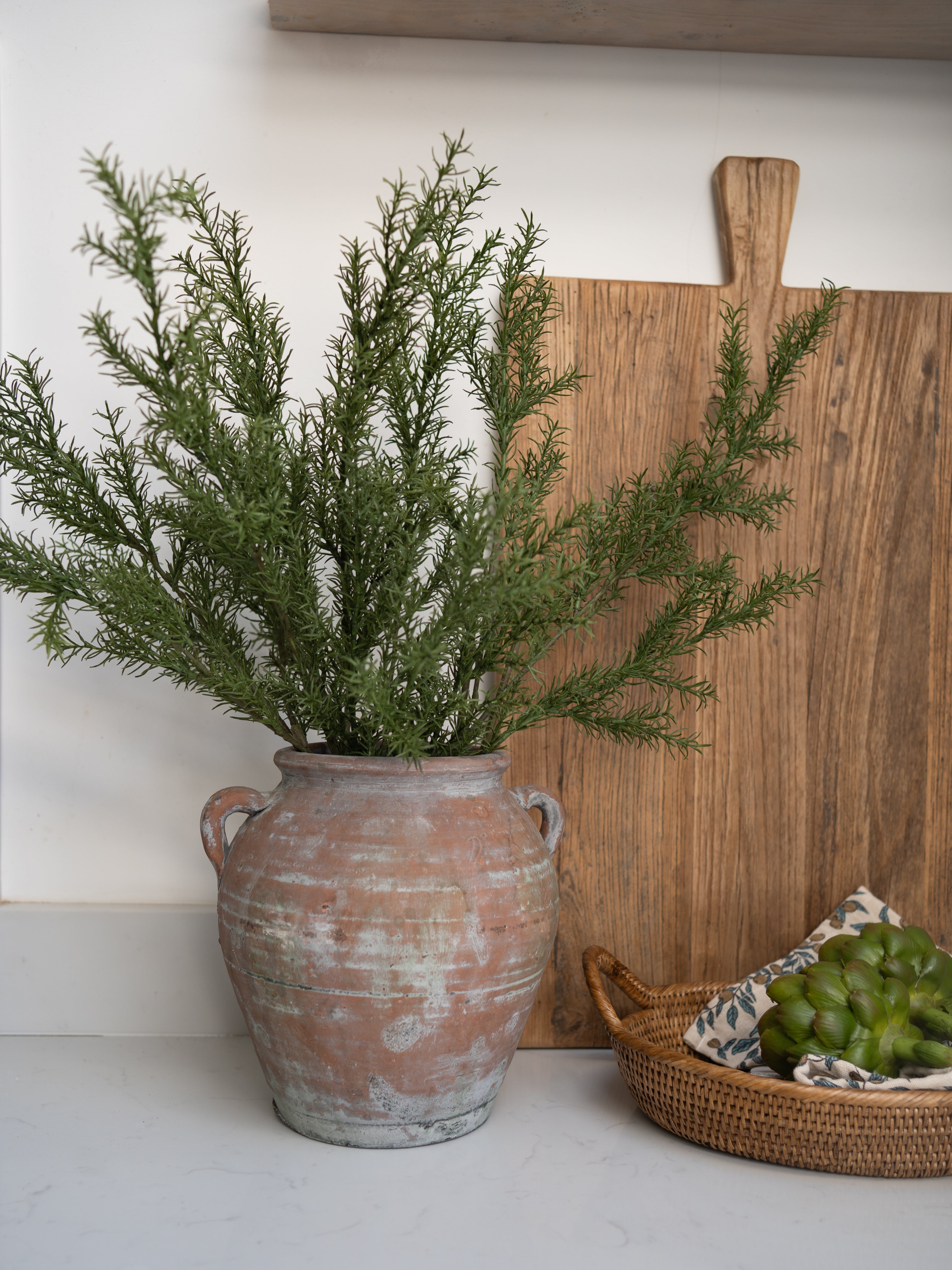faux rosemary stems in a terracotta olive pot with a wooden cutting board and basket on a white surface.