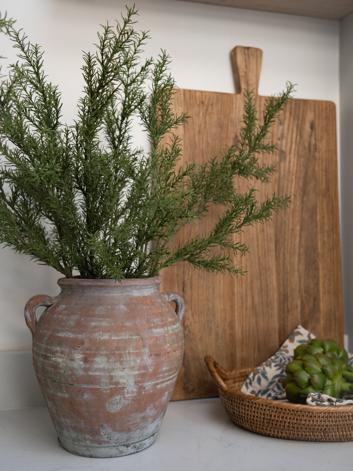 faux rosemary stems in a rustic terracotta pot with a wooden cutting board and woven basket in the background.