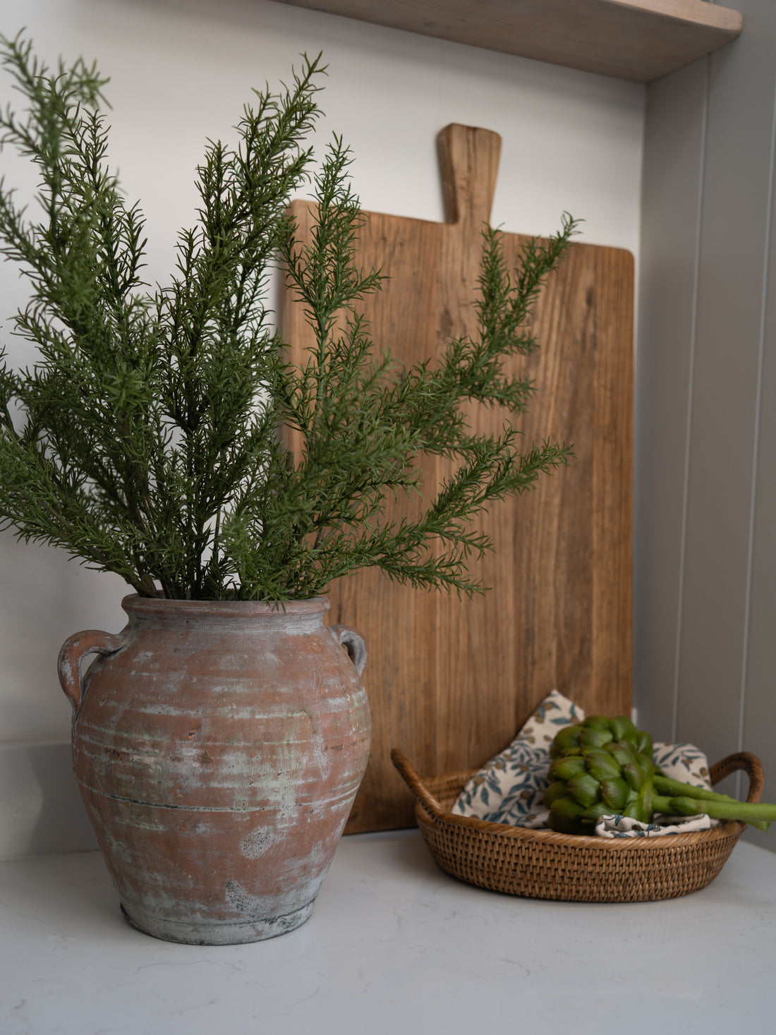 Rustic olive pot with faux rosemary  greenery, wooden cutting board, and woven basket on a white surface.