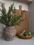 Rustic olive pot with faux rosemary  greenery, wooden cutting board, and woven basket on a white surface.