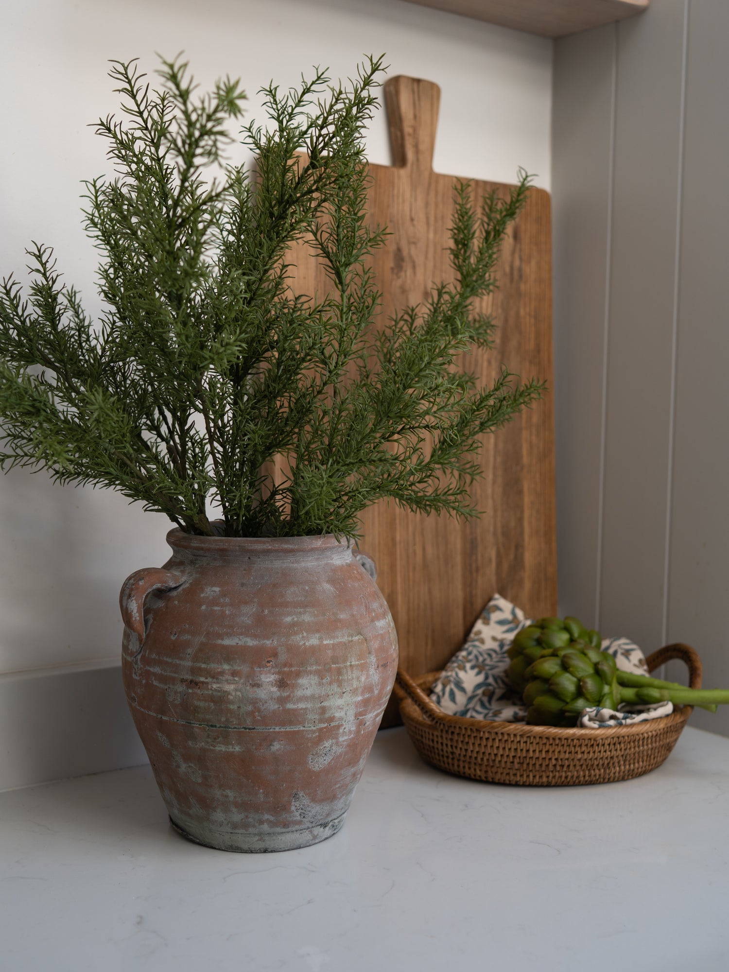 Terracotta pot with faux rosemary herb greenery on a white surface with a wooden cutting board and basket in the background.