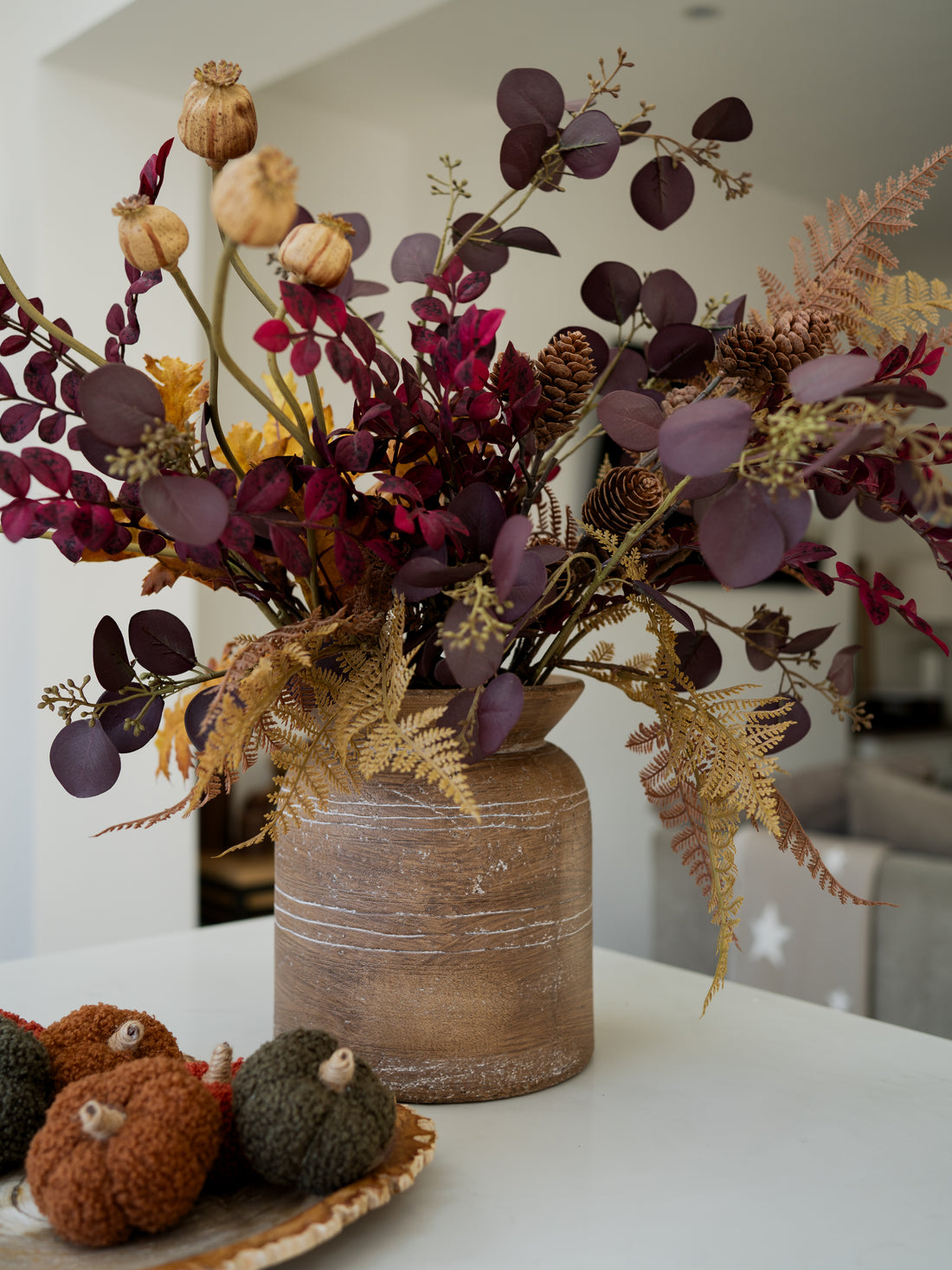 A rustic brown ceramic vase filled with a rustic autumn floral arrangement featuring burgundy eucalyptus leaves, tan seed pods, gold ferns, and pinecones, displayed on a white table in a modern interior setting.