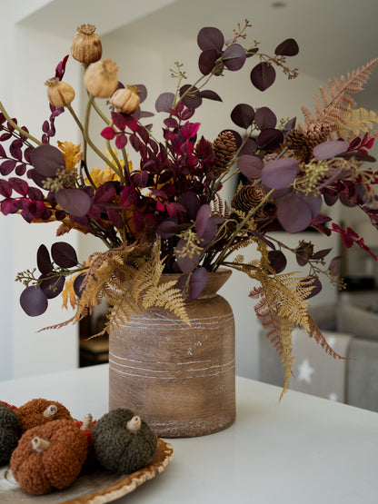 A rustic brown ceramic vase filled with a rustic autumn floral arrangement featuring burgundy eucalyptus leaves, tan seed pods, gold ferns, and pinecones, displayed on a white table in a modern interior setting.
