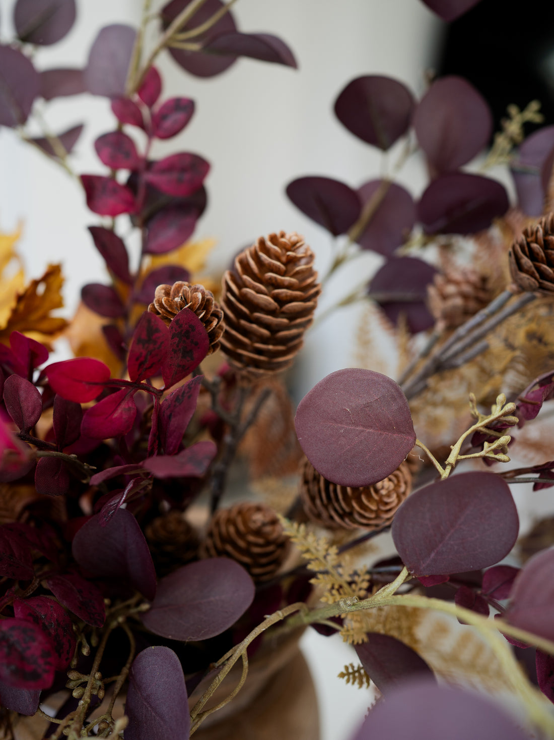 A close-up of a faux autumn floral arrangement featuring deep burgundy and plum leaves, natural brown pinecones, and tan grasses against a soft white background.