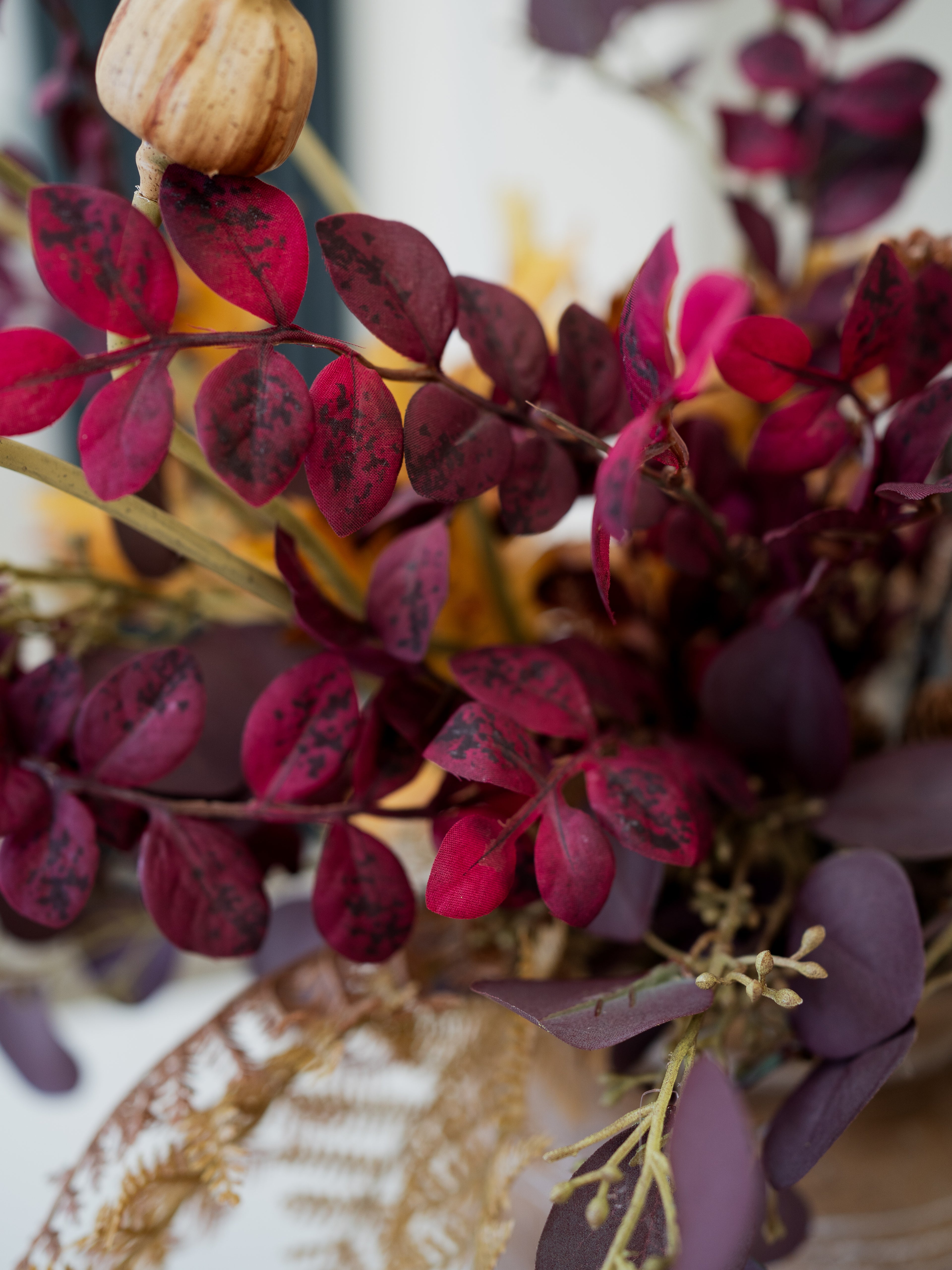 Close-up of an artificial autumn floral arrangement with deep burgundy and purple leaves, dried ferns, and a small beige decorative poppy on a light background.