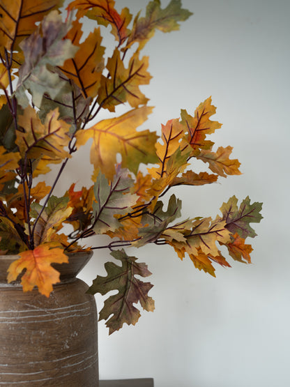 Autumn oak leaf artificial floral arrangement in a rustic brown ceramic vase against a plain light gray wall.