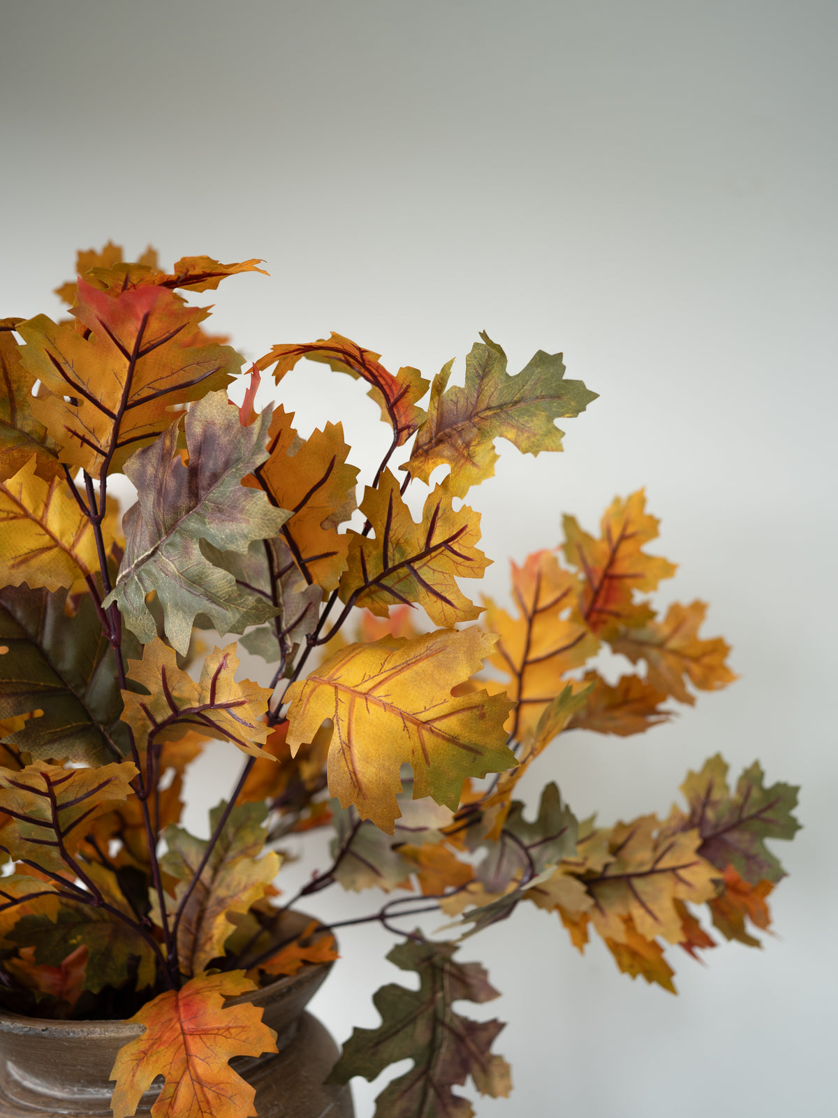 Autumn oak leaf artificial floral arrangement in a rustic ceramic pot, featuring yellow, orange, and green leaves with dark brown stems, set against a plain light gray background.