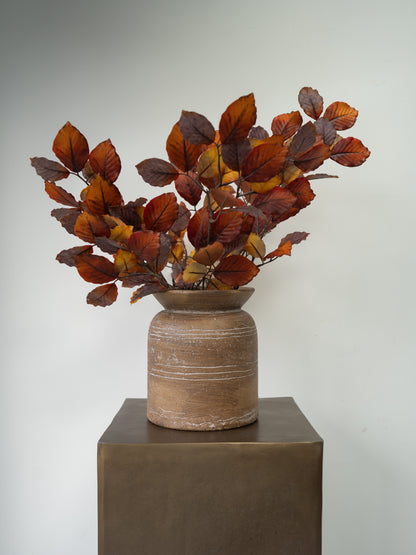 Autumn-colored artificial foliage with reddish-brown and orange leaves arranged in a rustic brown ceramic pot, displayed on a dark pedestal against a neutral gray background.