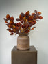 Autumn-colored artificial foliage with reddish-brown and orange leaves arranged in a rustic brown ceramic pot, displayed on a dark pedestal against a neutral gray background.