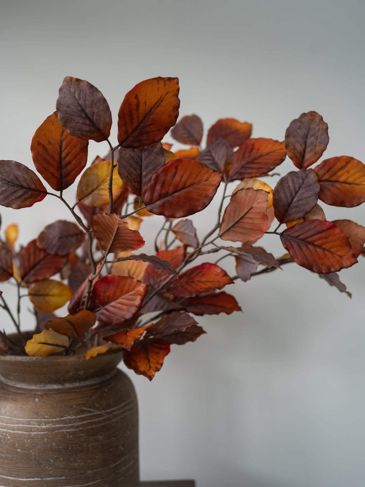 Autumn-colored artificial leaves in shades of orange, brown, and yellow, arranged in a rustic brown ceramic pot against a soft gray background.